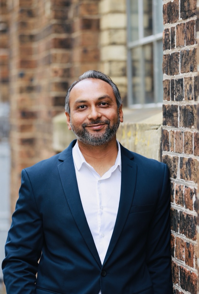 Anit Patel smiling, leaning against a brick wall